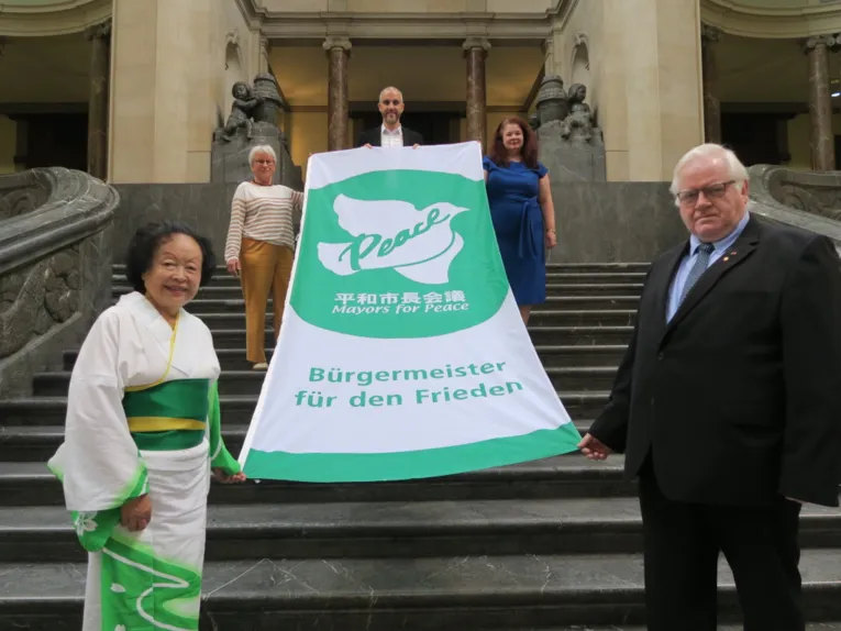 Oberb&uuml;rgermeister Belit Onay steht mit einem Mann und drei Frauen auf einer Treppe innerhalb des Neuen Rathauses in Hannover. Sie halten die Mayors-for-Peace-Flagge in den H&auml;nden.